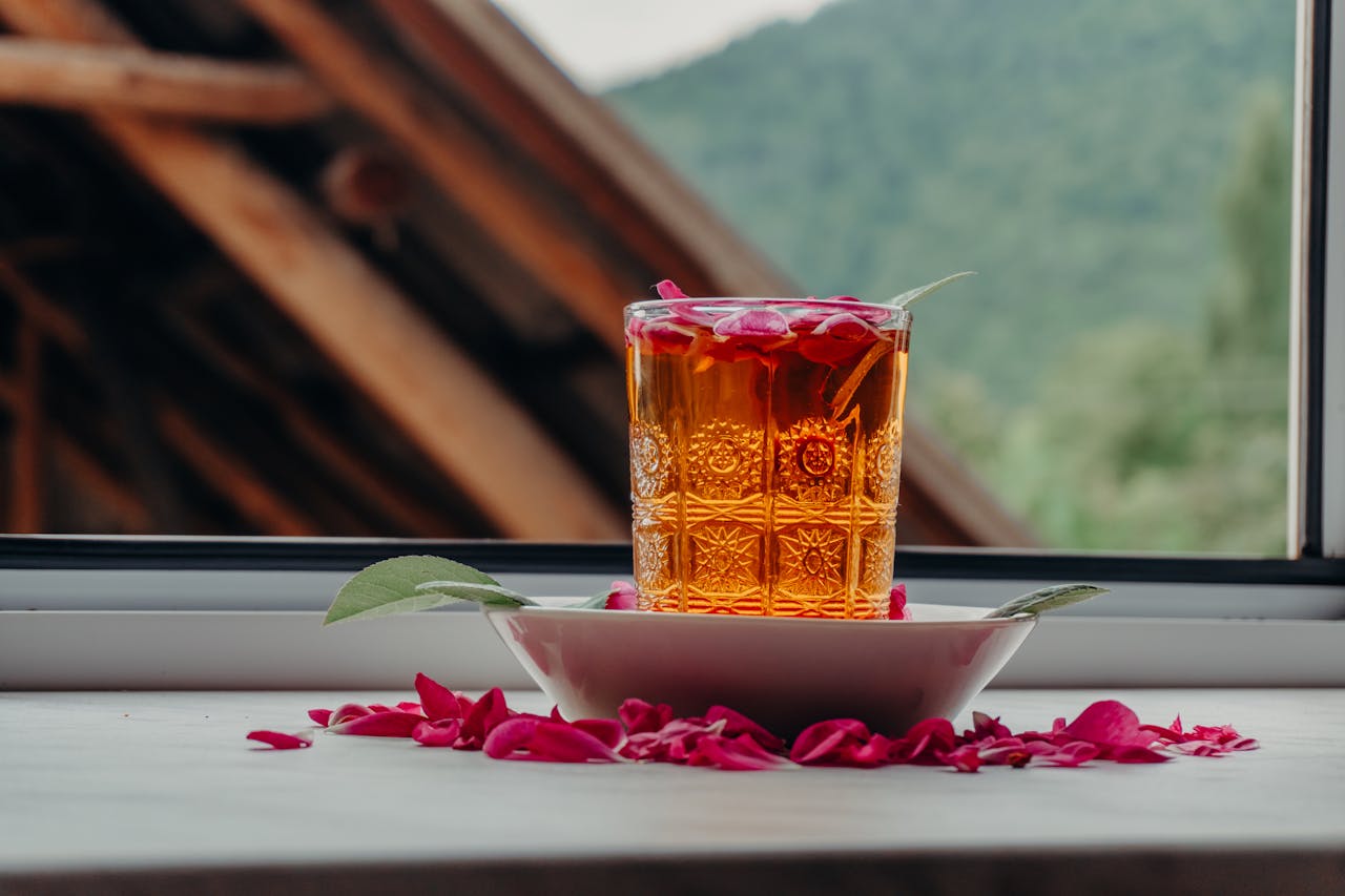 A glass of herbal tea garnished with rose petals and leaves, set on a windowsill with a scenic background.